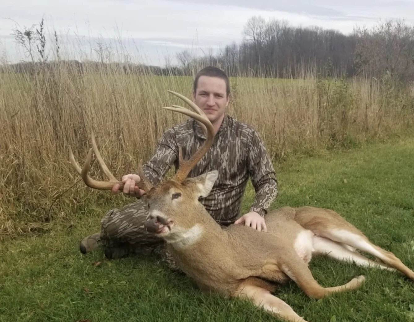 A man in camouflage clothing sitting on green grass, holding the antlers of a large deer lying on the ground, with a field and trees in the background.