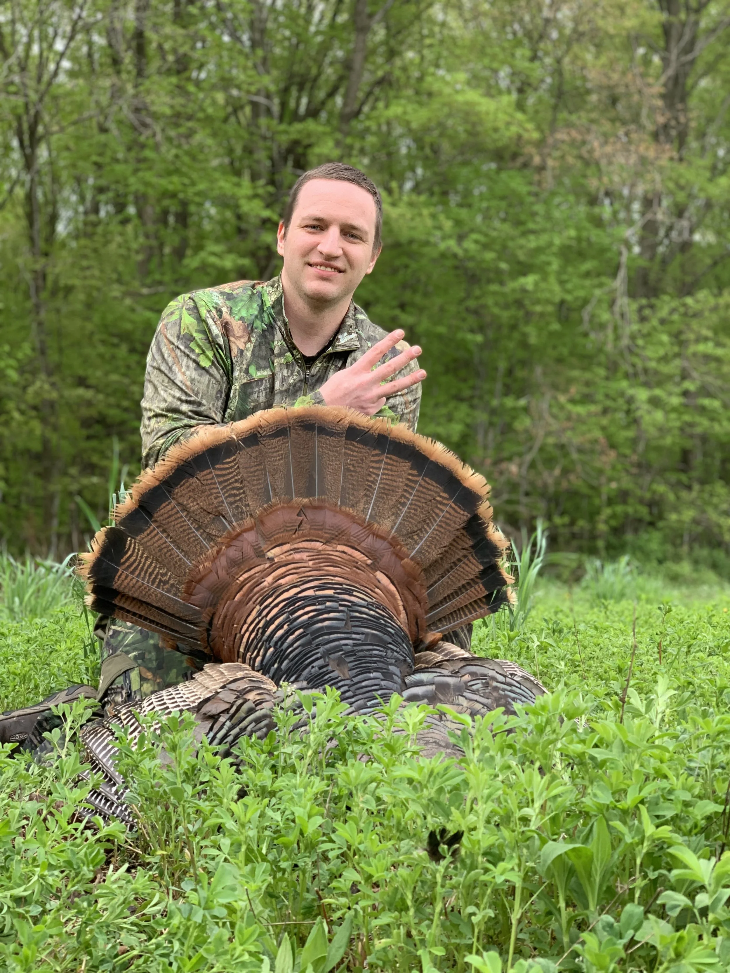 Man in camouflage clothing sitting in green field with tn turkey draped over his legs, forest background.