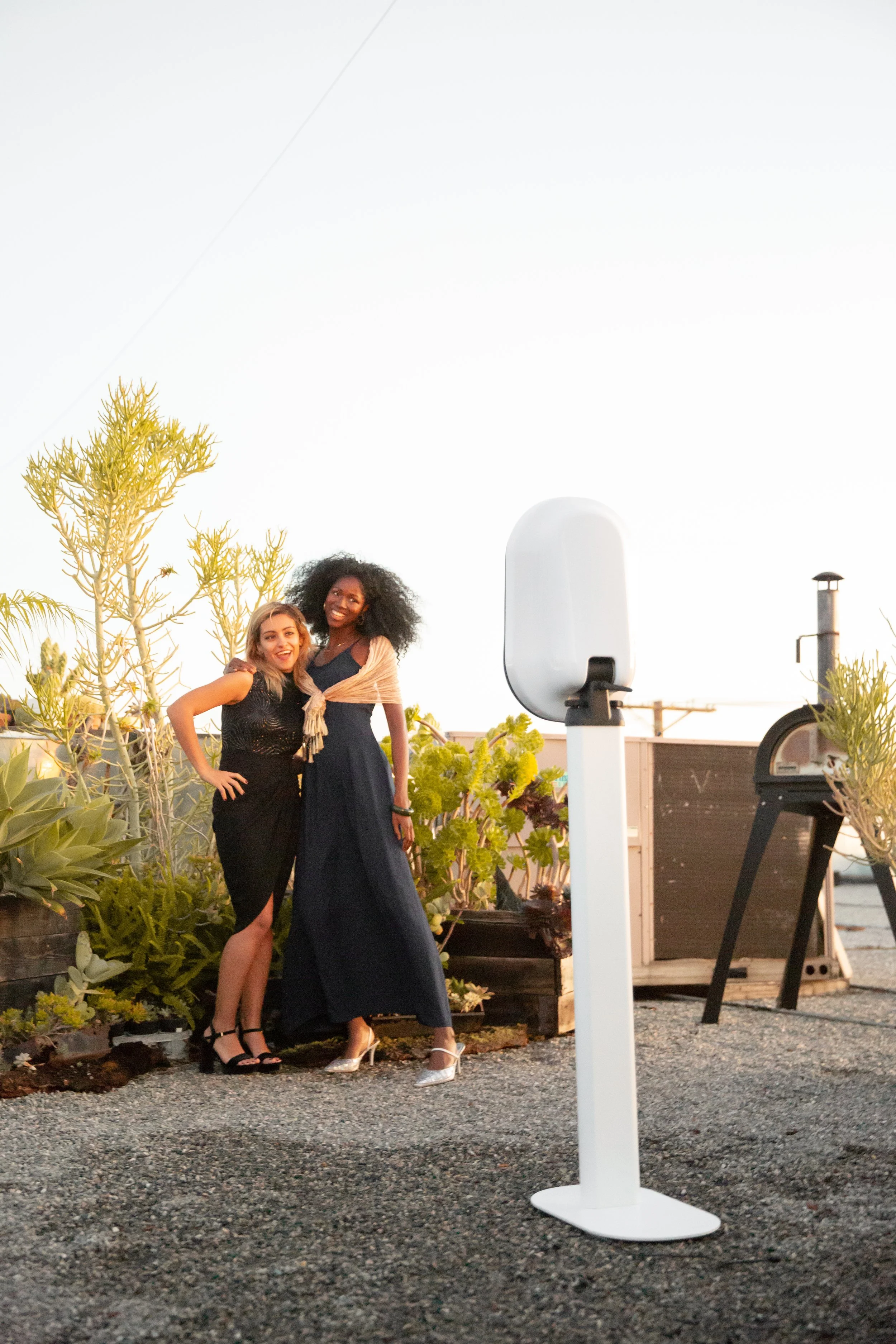 Two women dressed elegantly, smiling and posing together outdoors near some potted plants and garden furniture during sunset.
