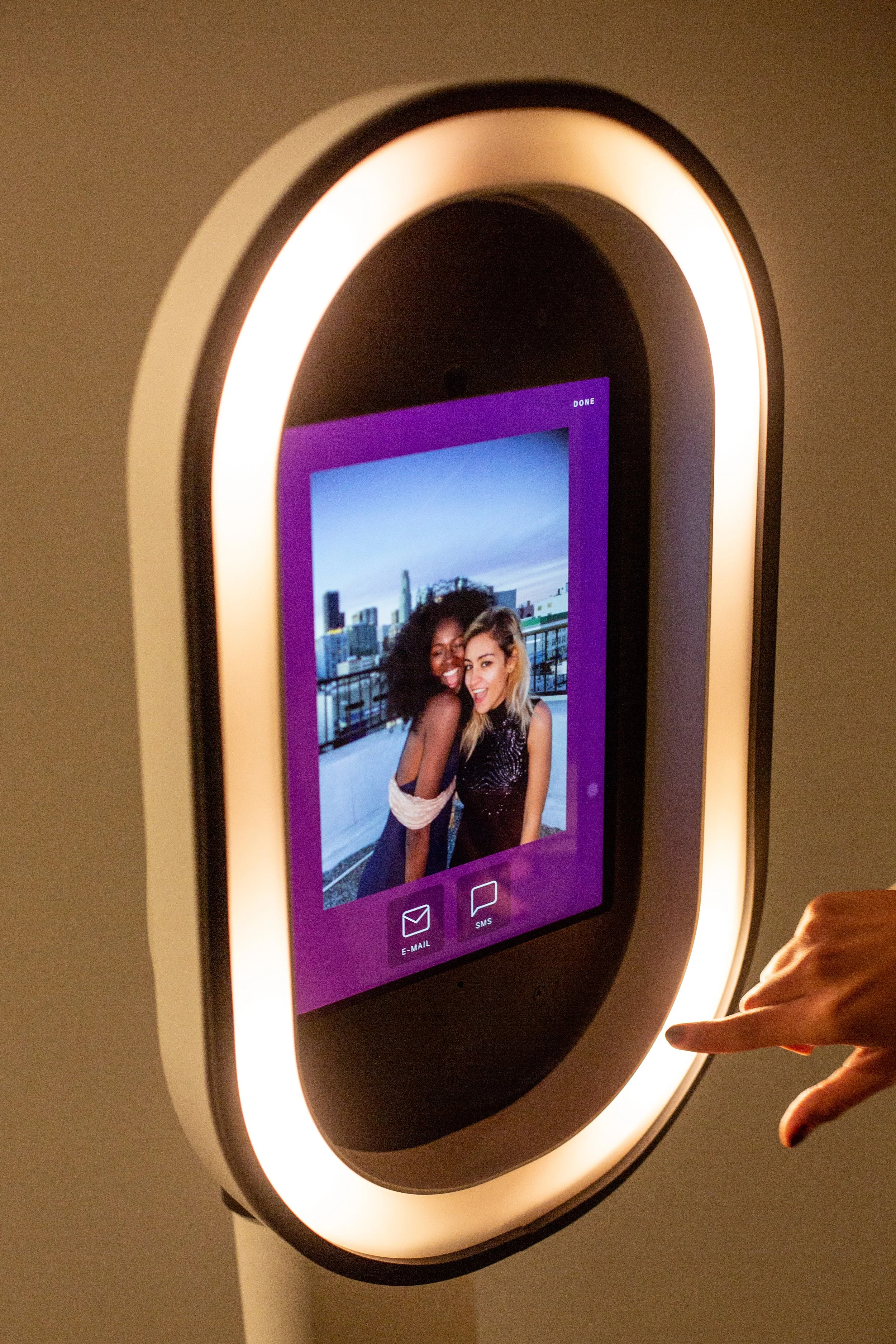 A touchscreen photo booth displaying a picture of two women smiling and posing together outdoors, with a city skyline in the background.