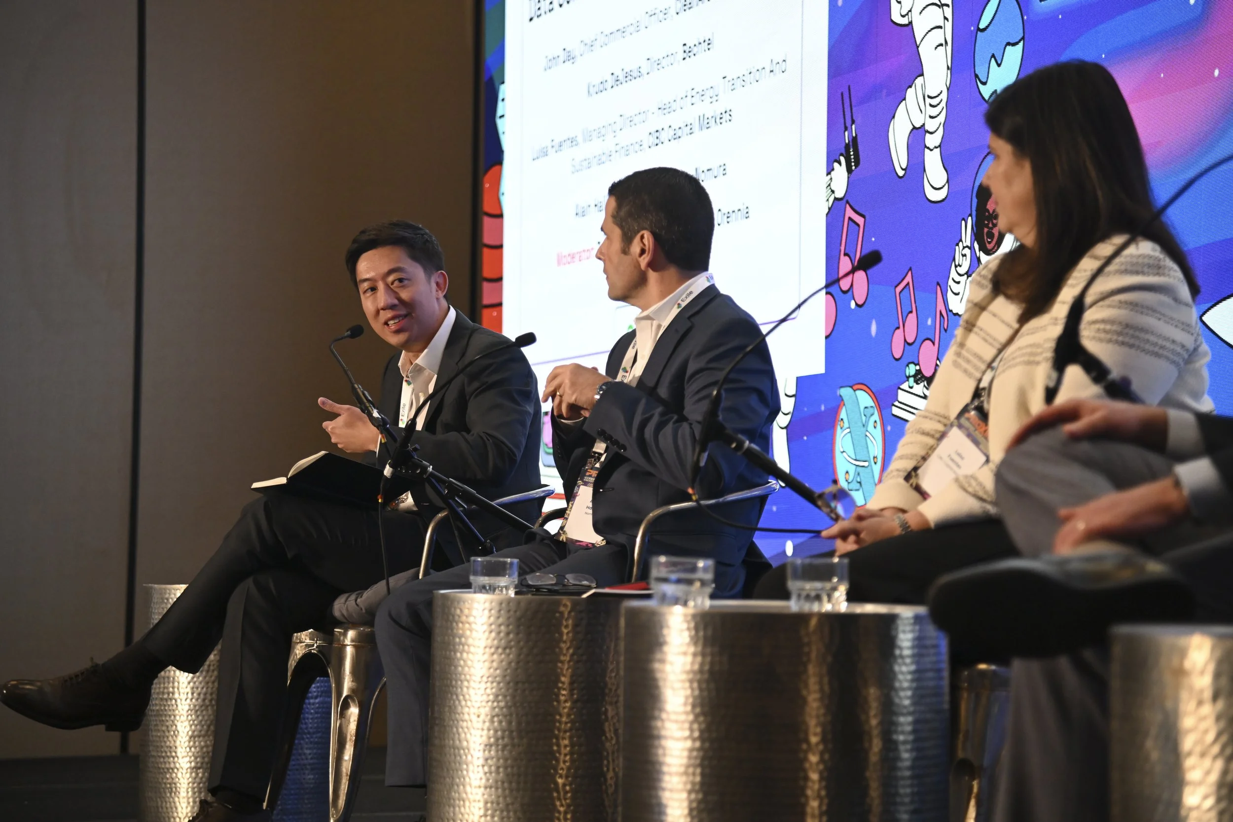 A panel of five individuals participating in a conference or panel discussion, sitting on stage with microphones, with a large screen in the background displaying text and colorful graphics.
