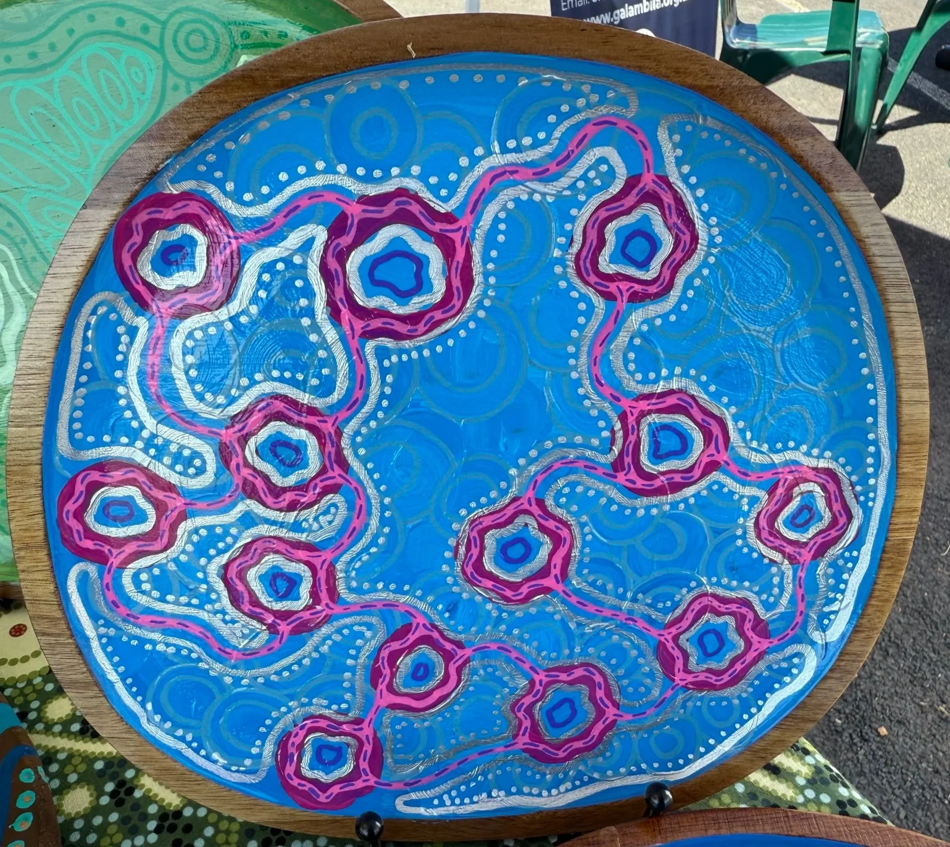 Colorful circular artwork with a blue background, pink and purple interconnected concentric shapes, and white dotted decorative patterns, displayed on a round wooden table.