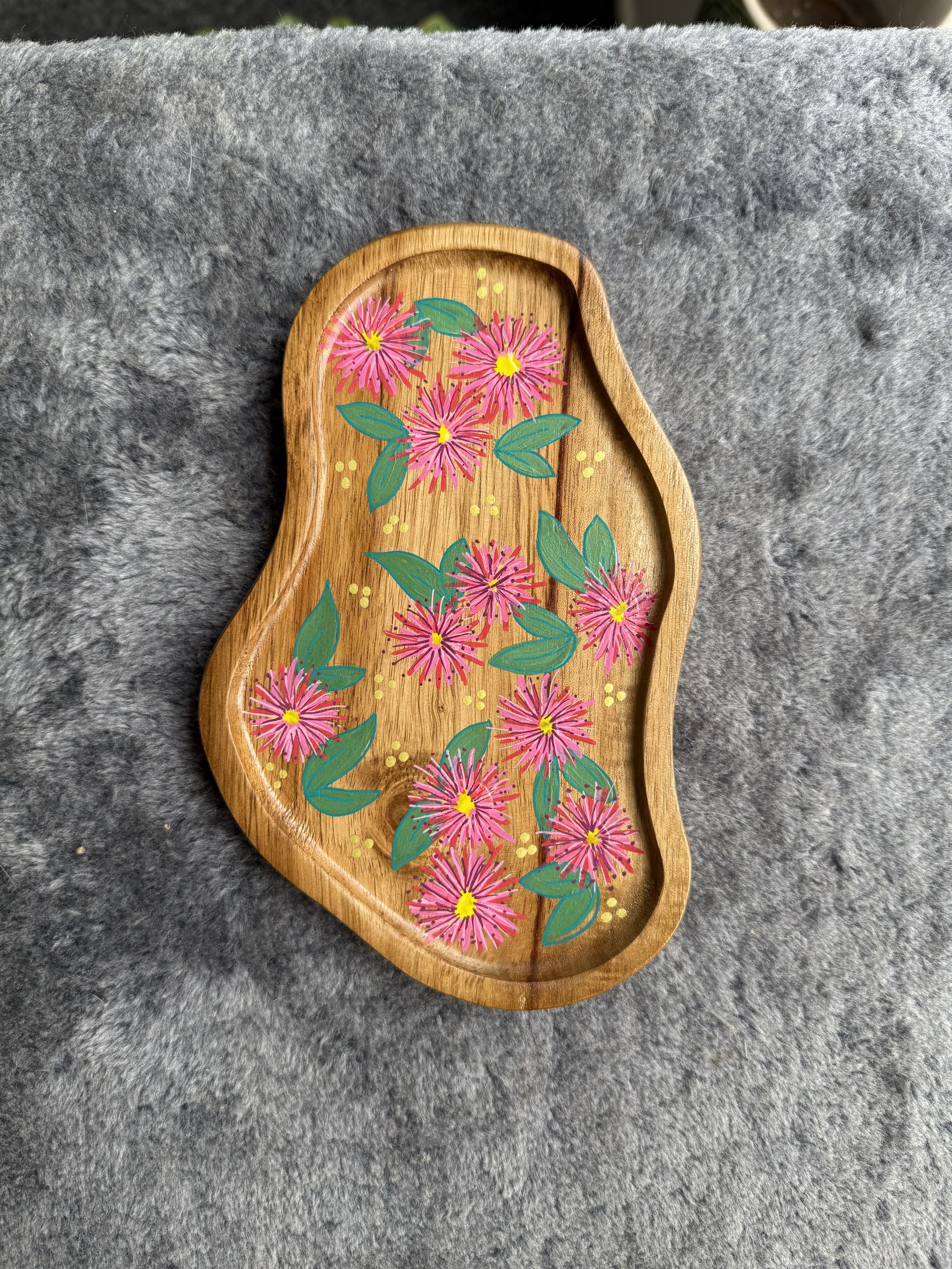 Wooden tray with a painted pink and yellow flower design with green leaves on a grey fabric surface.
