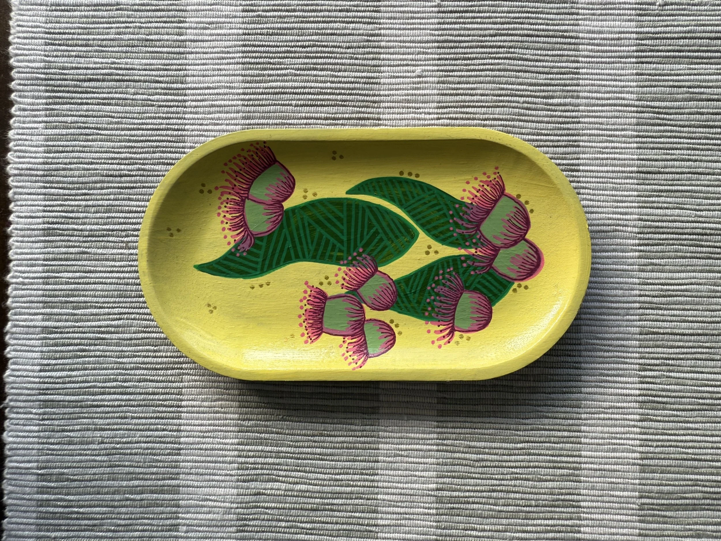 Yellow oval wooden tray decorated with pink and green floral design, placed on a gray and white striped fabric surface.