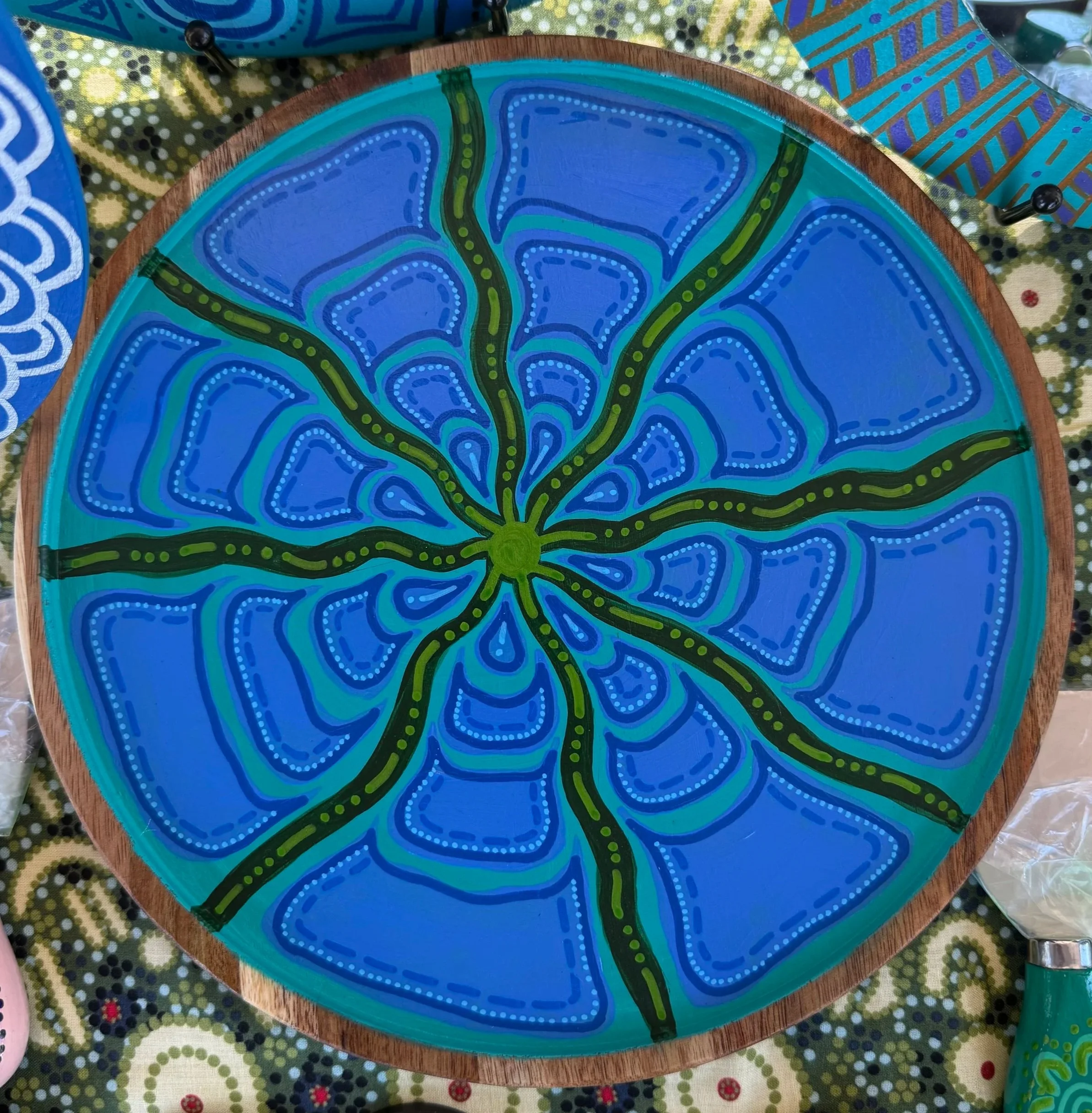 A round decorative wooden platter painted with a blue and green abstract design resembling a stylized flower or organic pattern.