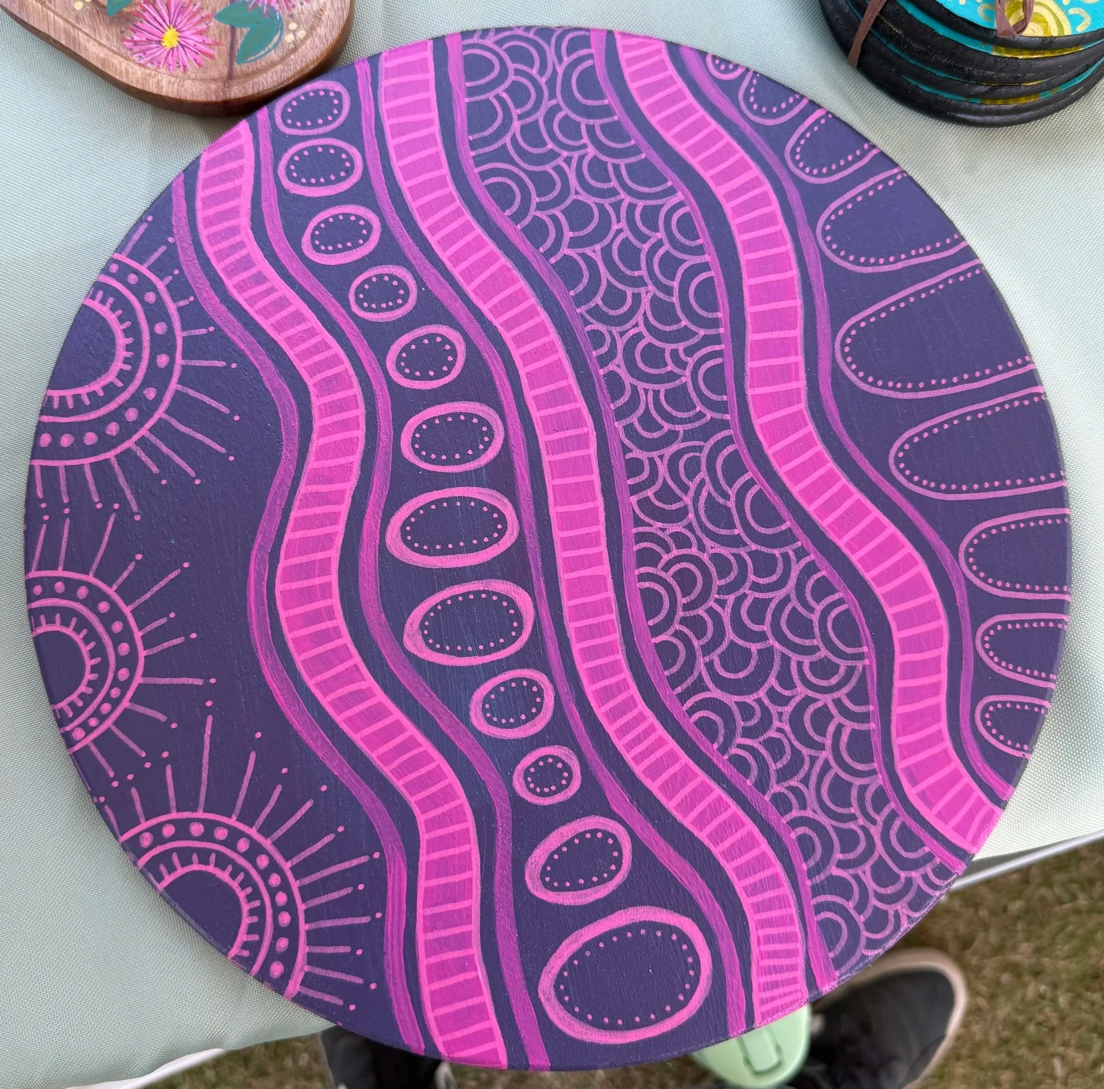 A round trivet with a vibrant, abstract painted design featuring bright pink lines and patterns on a dark purple background.