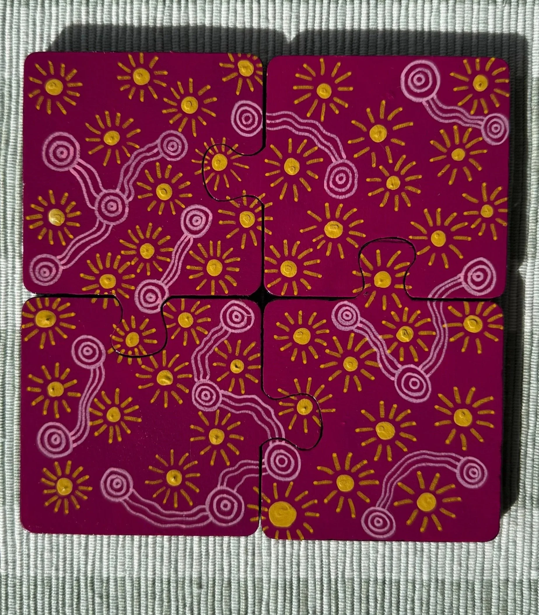 Four puzzle pieces with a pink background, yellow and white floral pattern, and interconnected white lines and circles, placed on a textured gray surface.