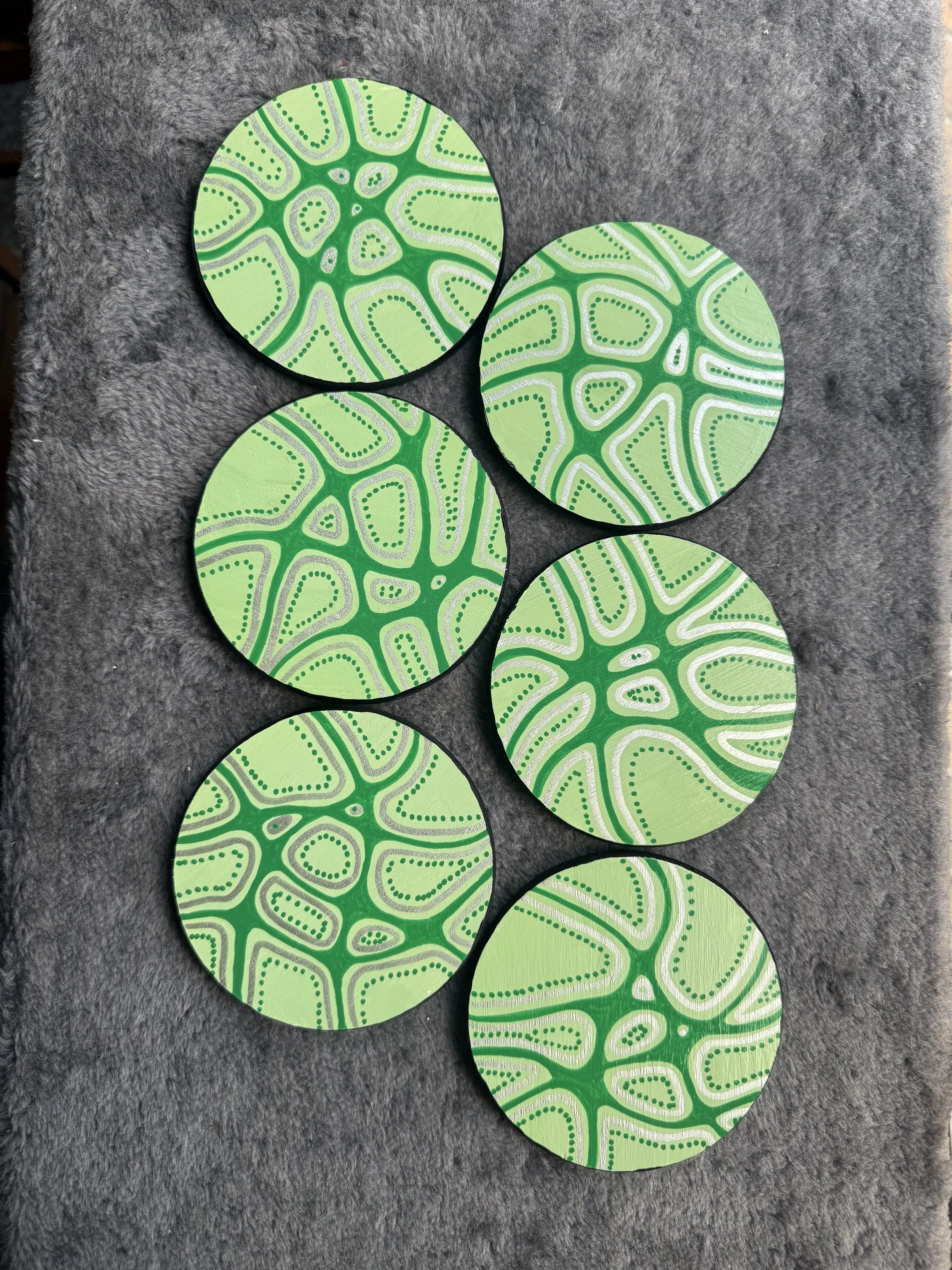 Five round painted coasters with green and white intricate abstract patterns, placed on gray fabric background.