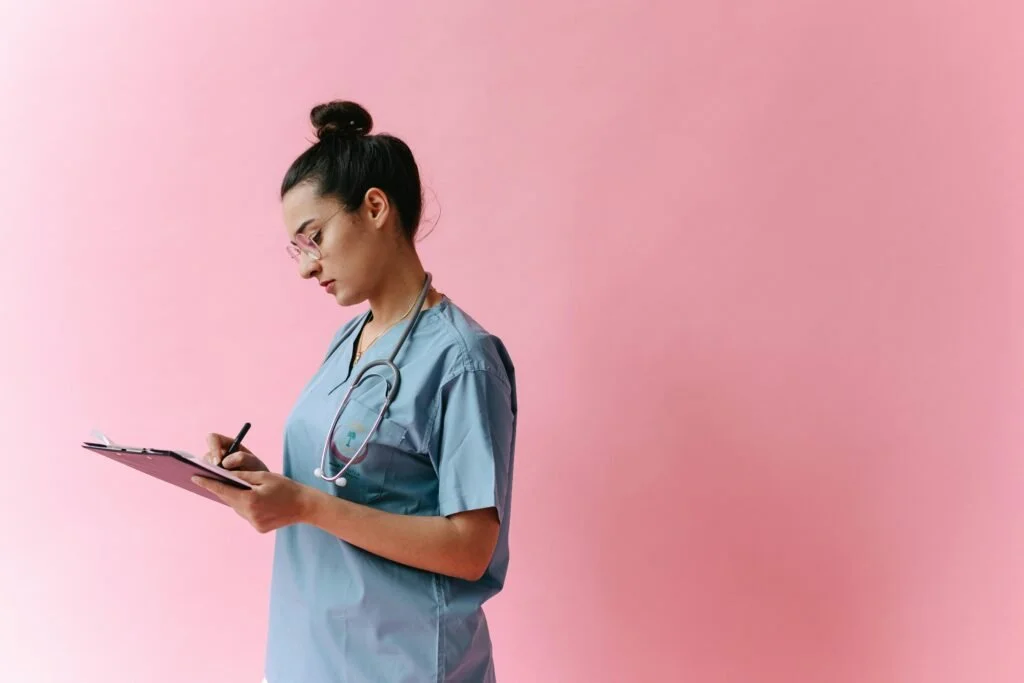 A female healthcare professional in scrubs with a stethoscope around her neck, writing on a clipboard against a pink background.