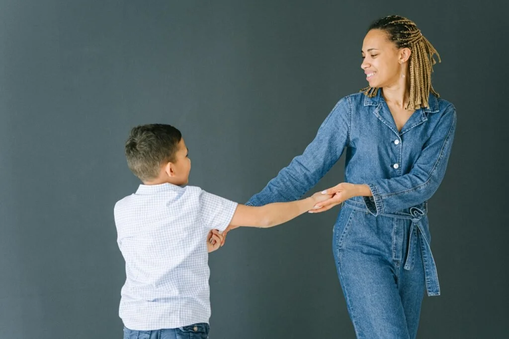 A woman with dreadlocks in denim outfit playing with a young boy against a gray background.
