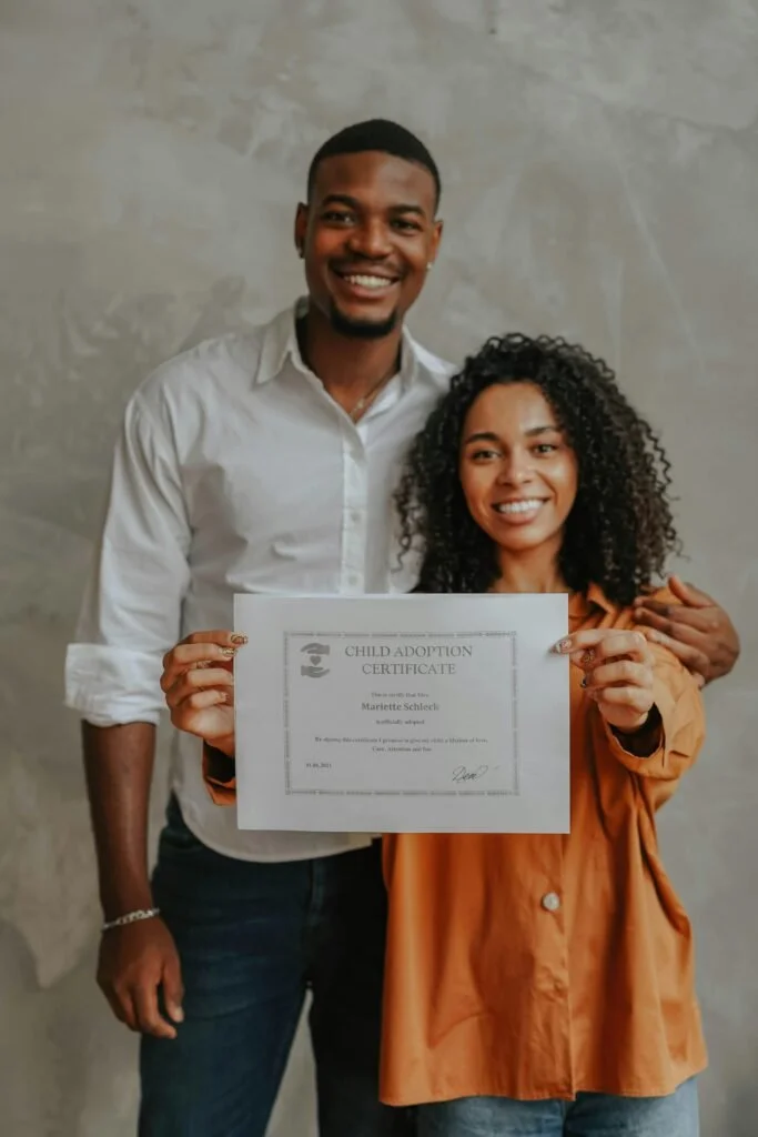 A smiling couple holding a child adoption certificate, with the woman in an orange jacket and the man in a white shirt, standing in front of a plain wall.