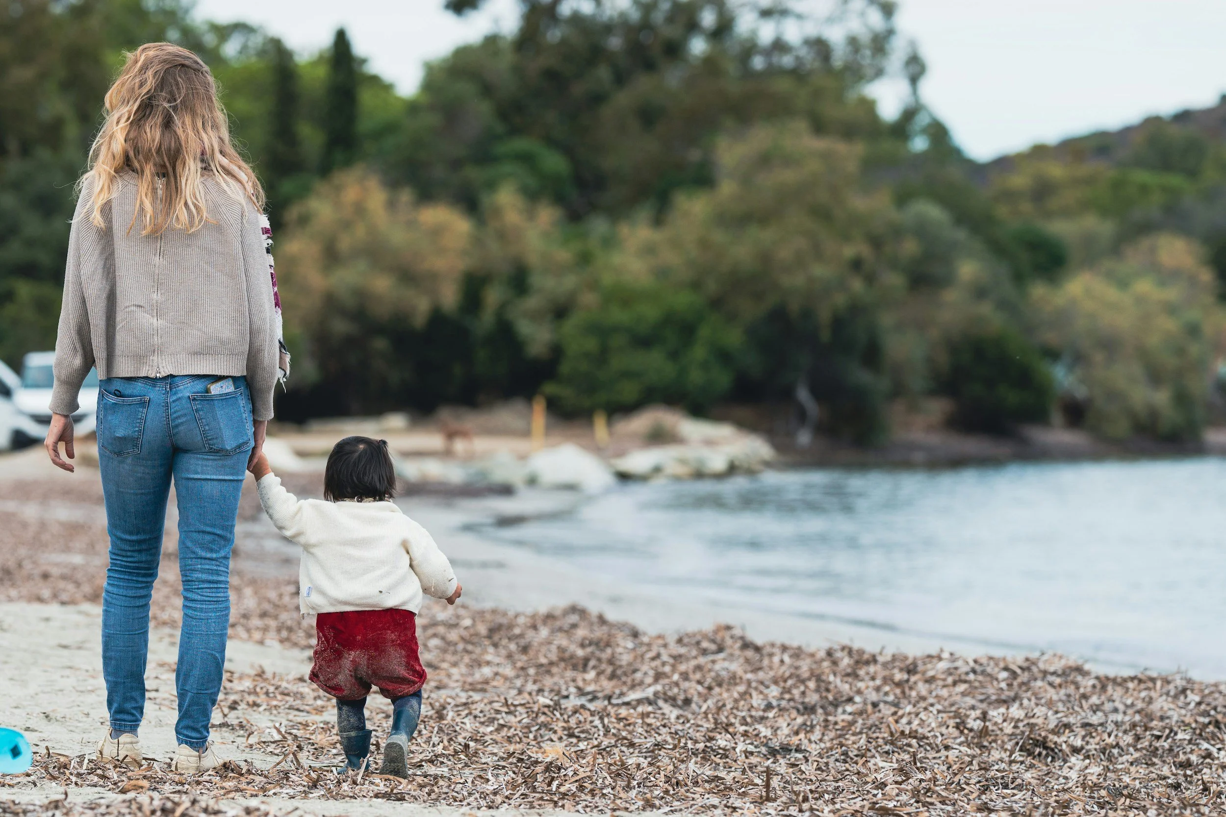 A woman and a young child walking hand in hand on a pebble-covered beach near a body of water, with trees in the background.