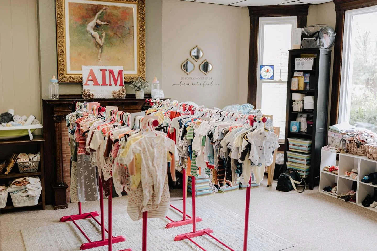 Multiple racks of baby clothes in a room with a large painting, decorative mirrors, and bookshelf.