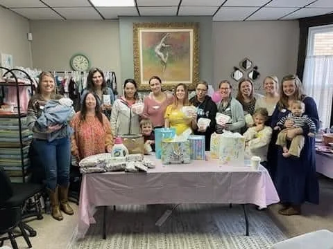 Group of women and children gathered around a table with gifts, in a room with a clothing rack, artwork, and decorative wall accents.