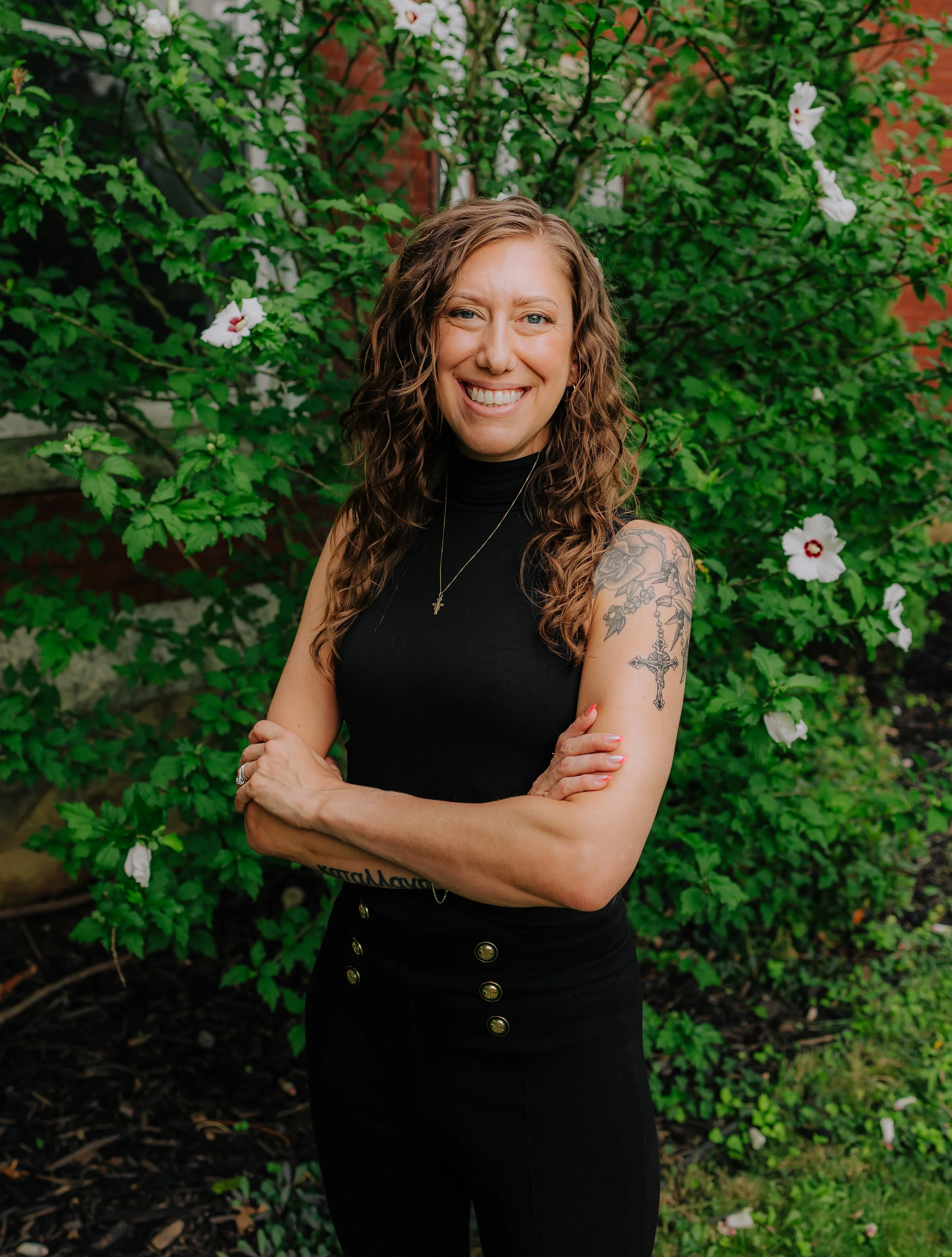 A smiling woman with curly hair and tattoos on her arm, standing in front of a green, flowering bush.