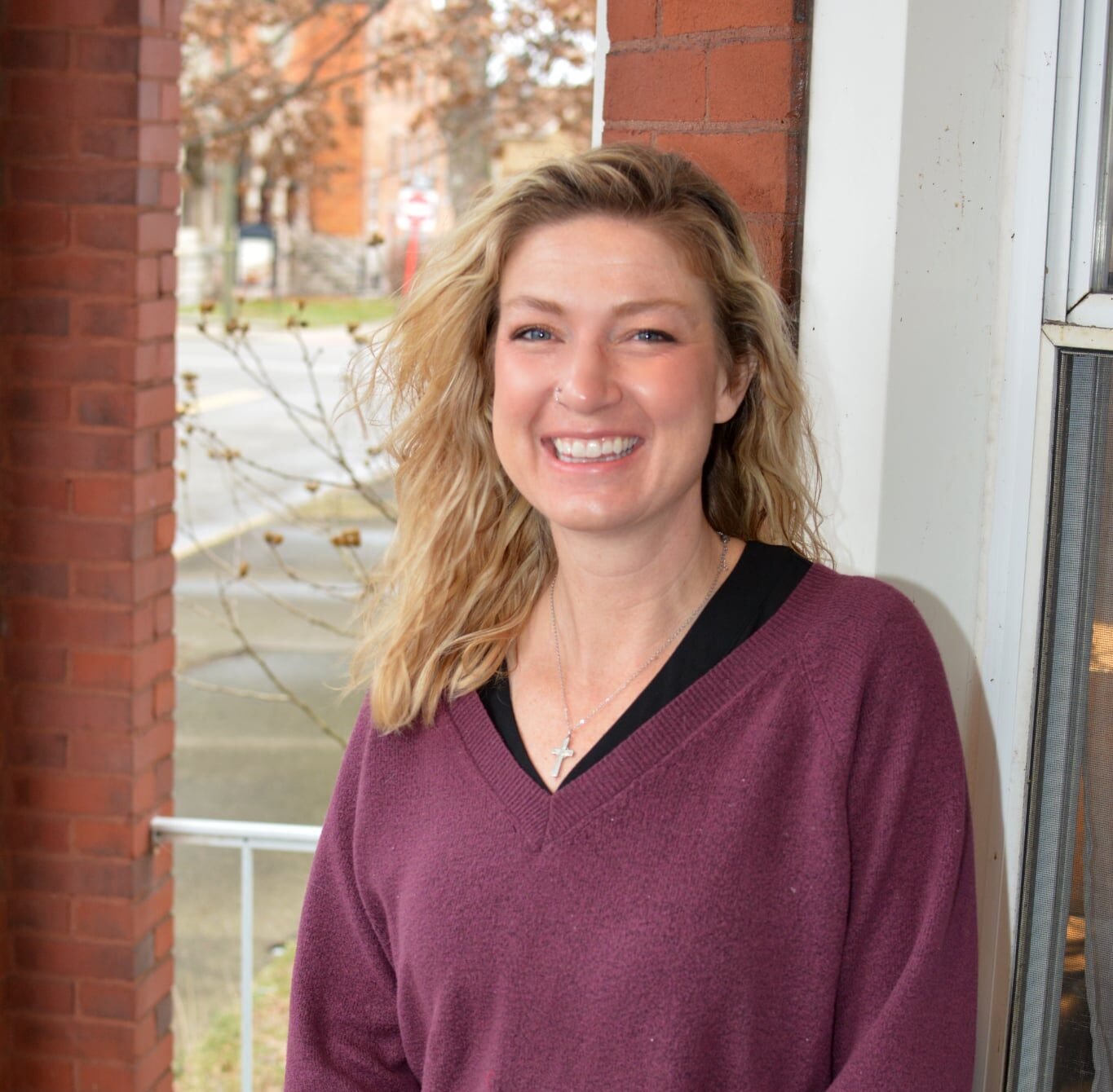 A smiling woman with blonde, wavy hair, wearing a purple sweater, standing next to a white wall and brick column, with a tree and street in the background.