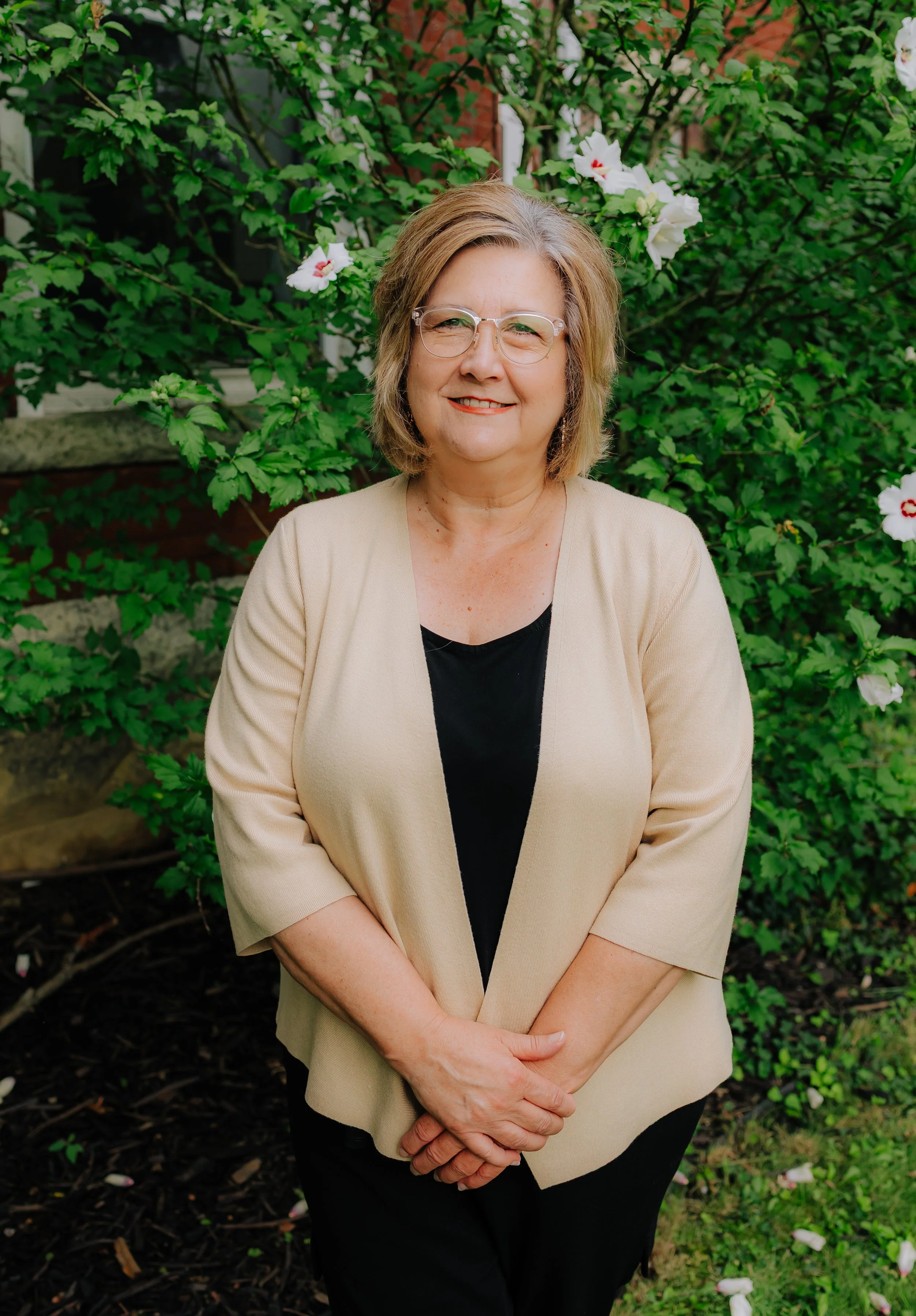 A woman with shoulder-length blonde hair, wearing glasses, a black top, and a beige jacket, standing outdoors in front of a bush with white flowers.
