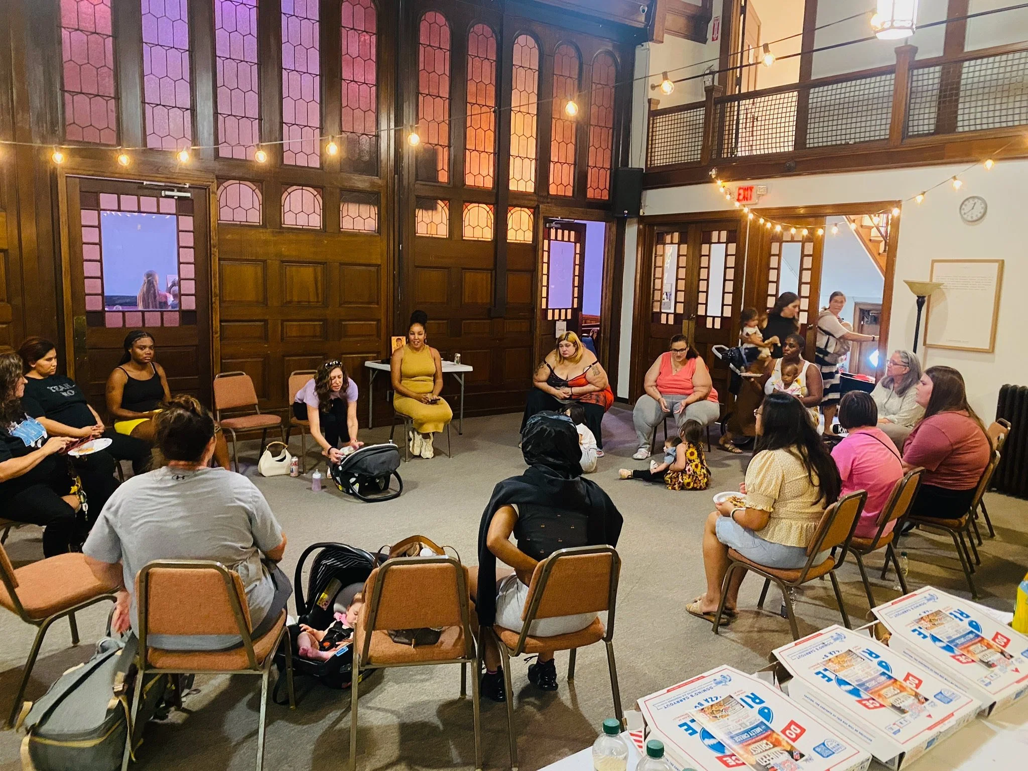 A group of diverse women and children sit in a circle inside a warmly lit, wood-paneled room with large windows. Some women are holding plates of food, and a woman in a yellow dress is speaking or leading the group. Pizza boxes are on a table in the foreground.