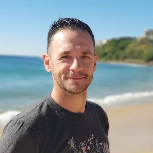 Man with dark hair and beard, smiling, standing on a beach with ocean and land in the background on a sunny day.