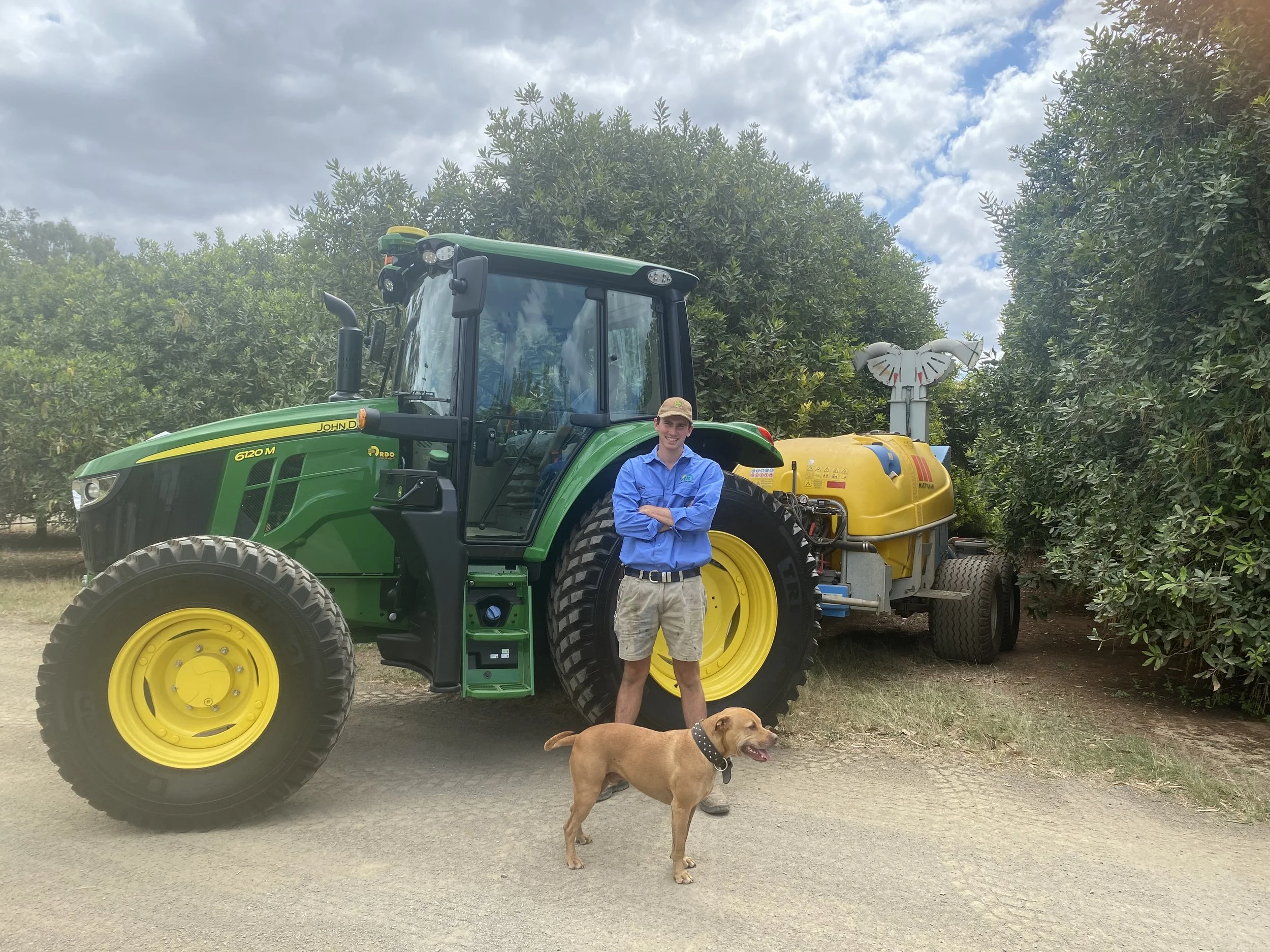A young man standing in front of a large John Deere tractor with a yellow spraying attachment, a brown dog with a black collar, and green trees in the background.