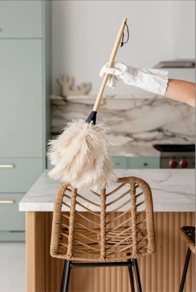 Person wearing white gloves holding a feather duster, cleaning a wicker chair in a modern kitchen.