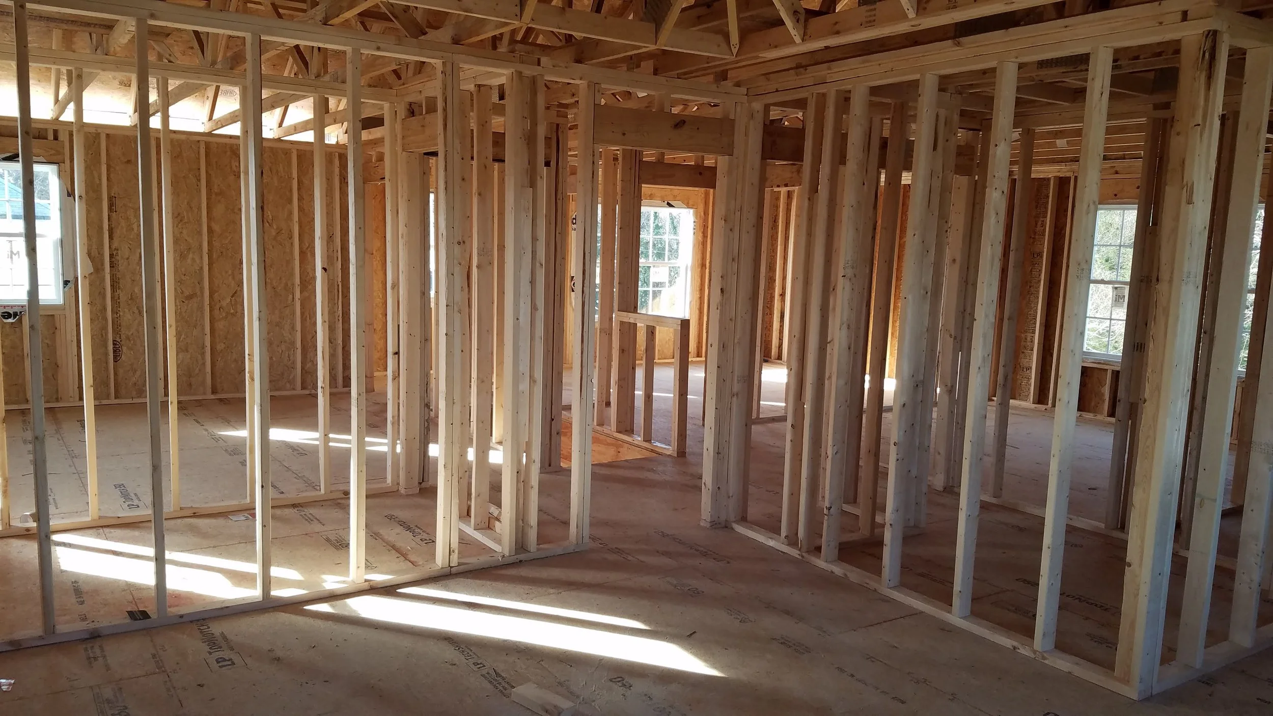 Interior of a house under construction with exposed wooden framing and unfinished floors, windows allowing natural light.
