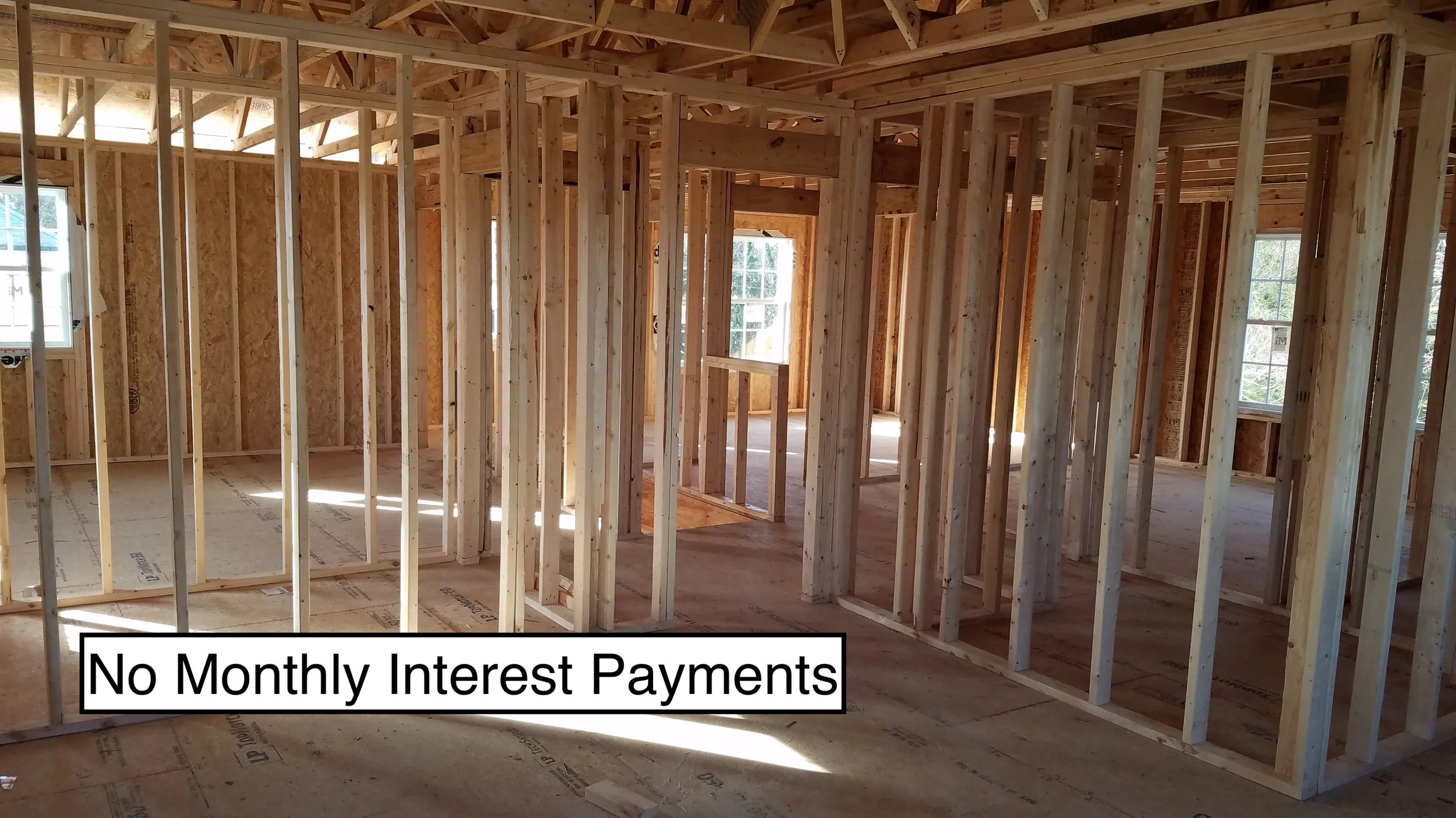 Interior of a house under construction with exposed wooden framing and plywood subfloor, sunlight coming through windows, and the caption "No Monthly Interest Payments".