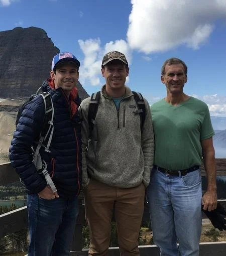Three men standing outdoors on a mountain overlook, with cliffs and sky in the background.