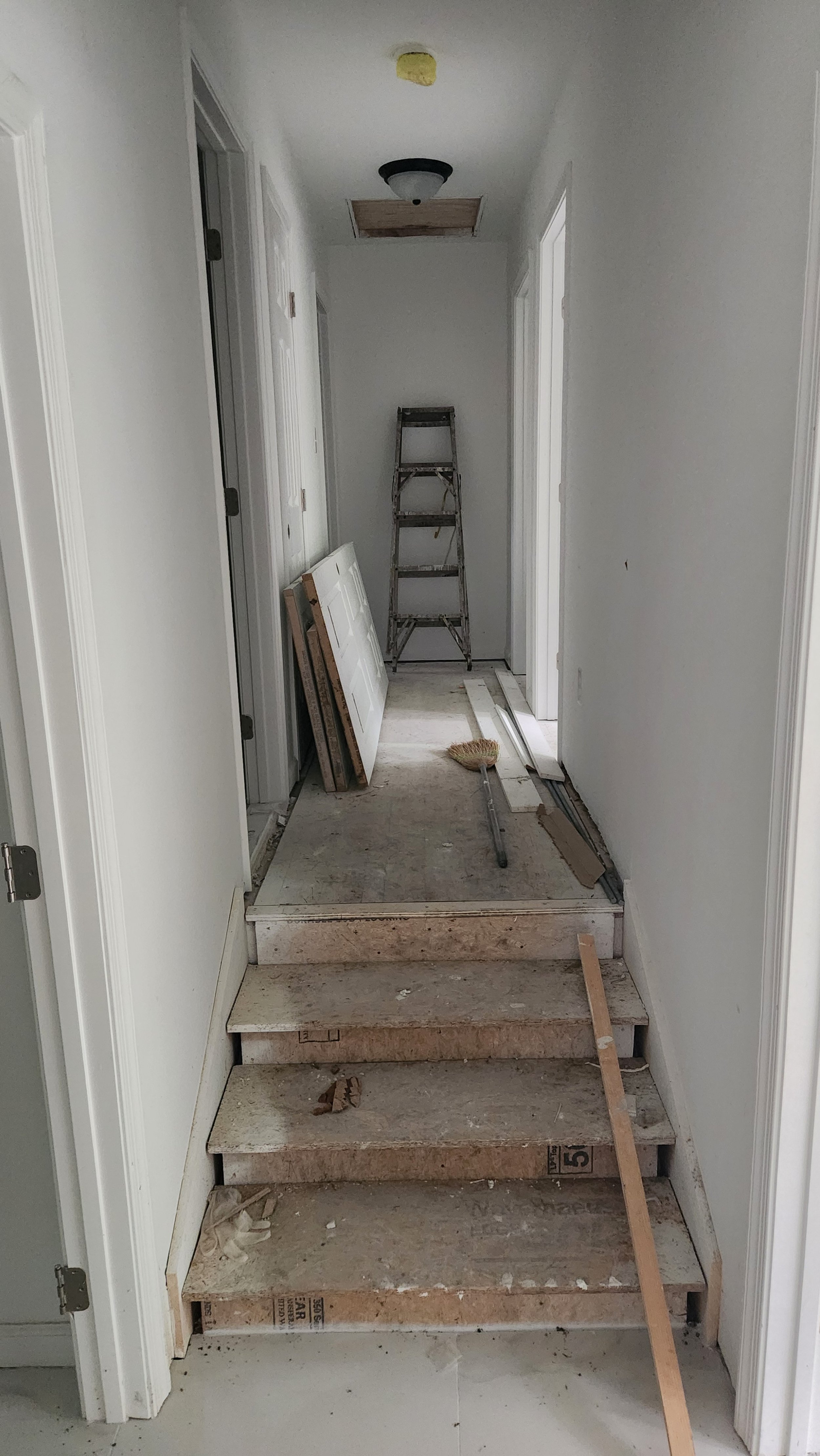 Interior hallway under renovation with exposed stairs, leaning door frame, ladder, construction tools, and materials.
