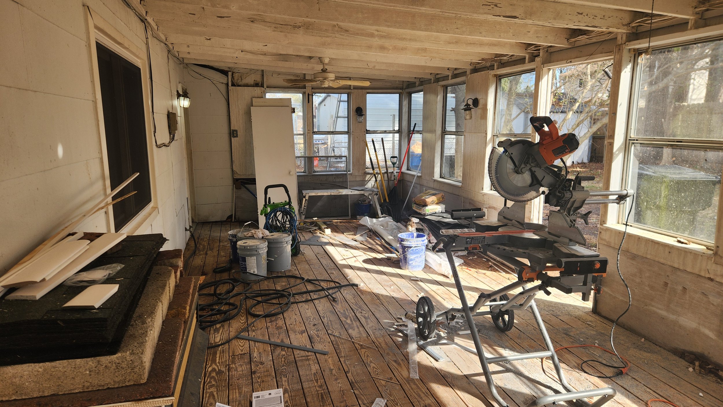 A sunlit enclosed porch or sunroom under renovation with construction tools including a large electric saw, paint buckets, various hand tools, and building materials scattered on the floor, with several windows letting in natural light..