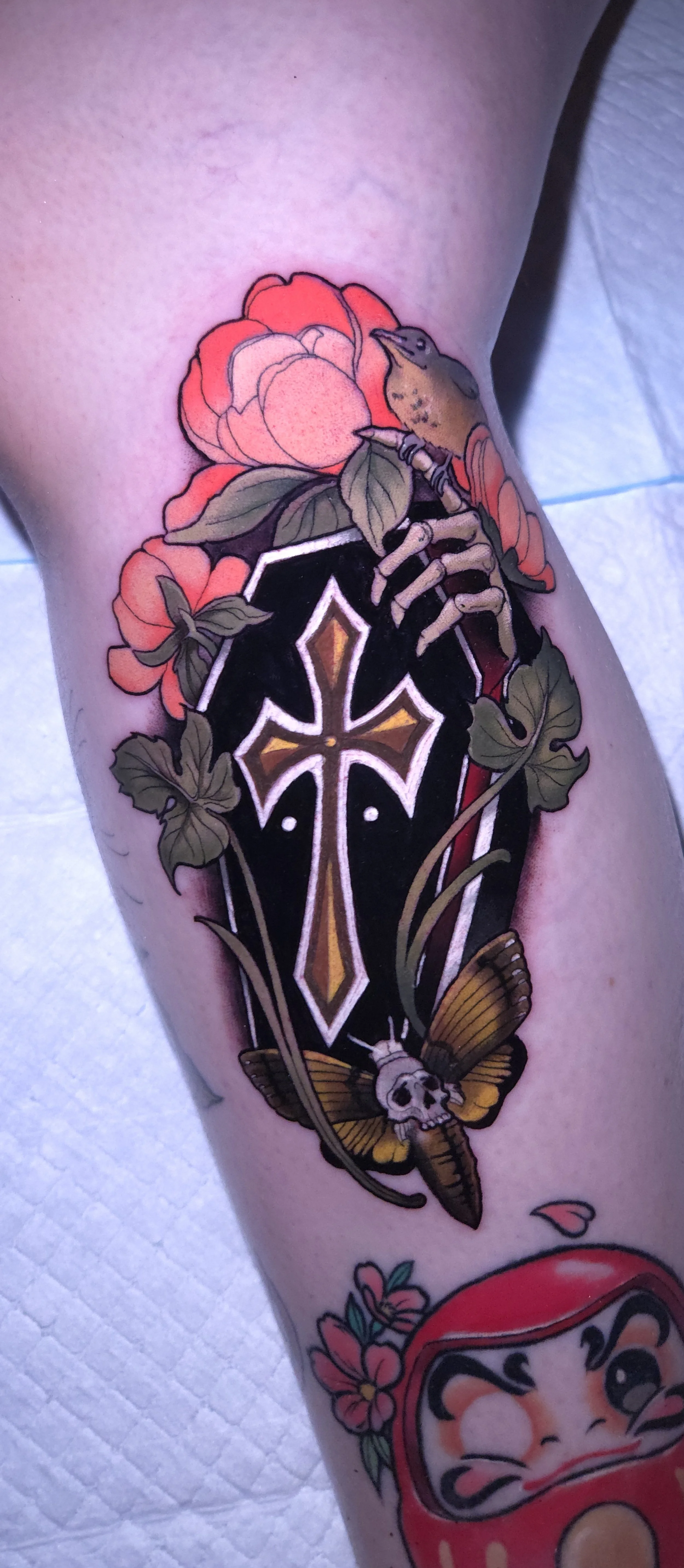 Tattoo featuring a colored cross with floral and botanical elements, a moth with a skull on its head, and a small bird, all outlined in bold black lines.