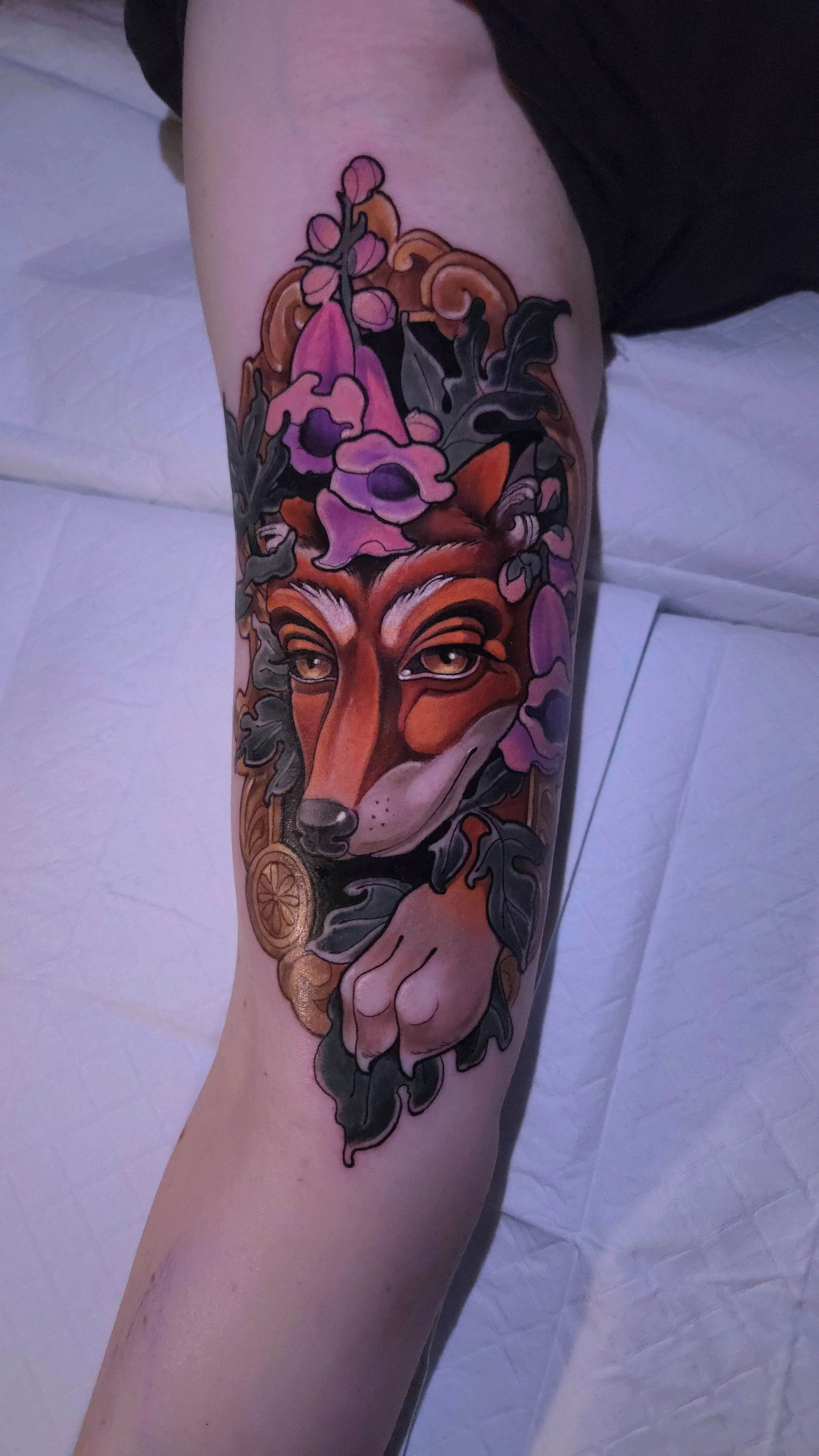 Tattoo of a fox's face with detailed eyes, surrounded by purple flowers and leaves, with a hint of a decorative element at the penguin's neck.