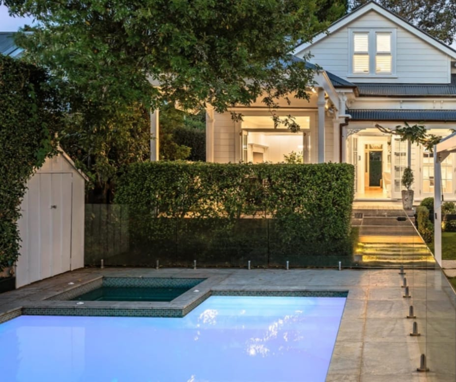 A backyard view of a house with a swimming pool in the foreground, hedges, trees, and a porch lit up at dusk.