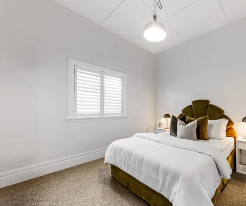 A bedroom with a white bed, multiple pillows, bedside lamps, a window with shutters, and a hanging ceiling light.