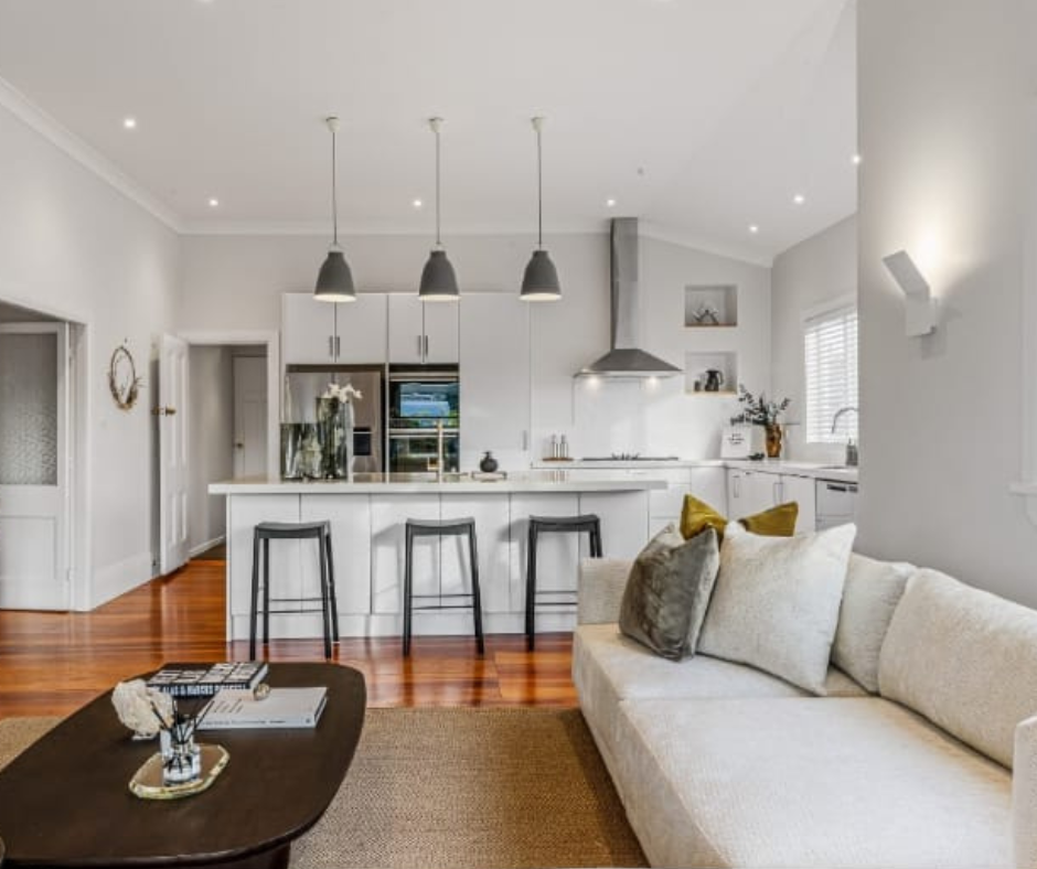 Modern, white kitchen and living room with a beige sofa, black bar stools, and wooden flooring.