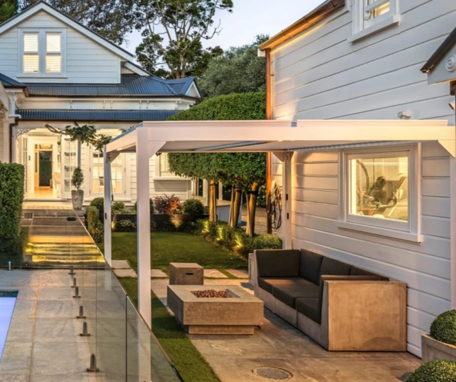 Exterior view of a modern house with a backyard patio, outdoor sofa, fire pit, glass railing, and landscaped garden during dusk.