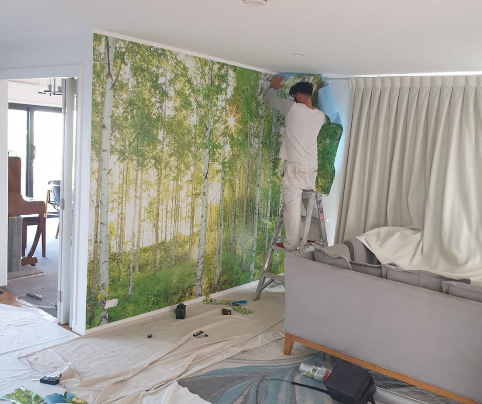 Person hanging wallpaper with a forest scene of green trees and sunlight in a living room, standing on a step stool, with tools and supplies scattered on the floor.
