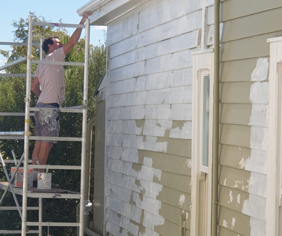 Person on scaffolding painting the exterior of a house white, with some areas already painted and some still needing paint.