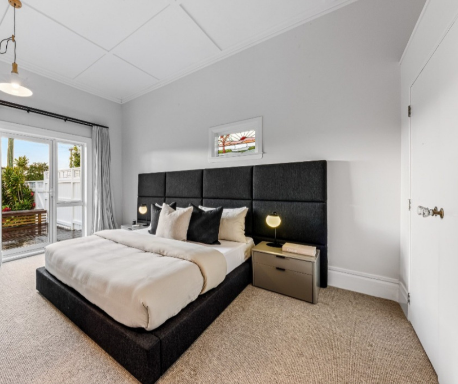 Modern bedroom with a black upholstered bed, white bedding, multiple pillows, nightstand with a lamp, small window above the bed, sliding glass door leading to an outdoor patio, and beige carpet.