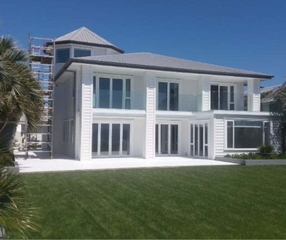 Modern white two-story house with large windows and a gray metal roof, surrounded by a green lawn and palm trees, under a clear blue sky.