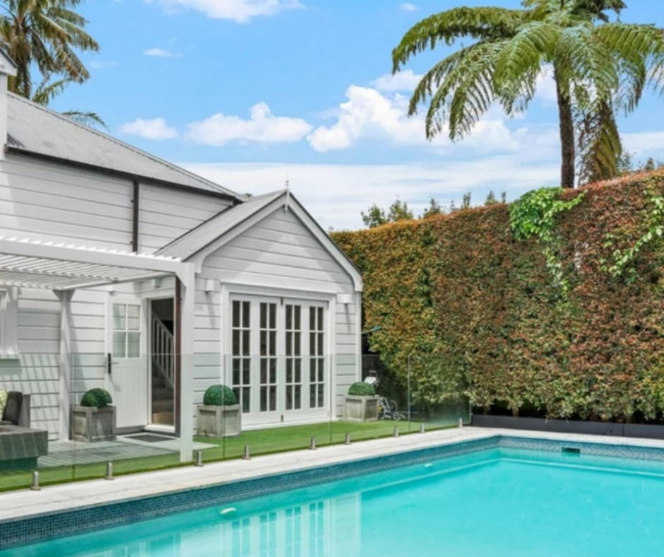 A backyard with a swimming pool, white house with large glass doors, and tall trees and bushes in the background.