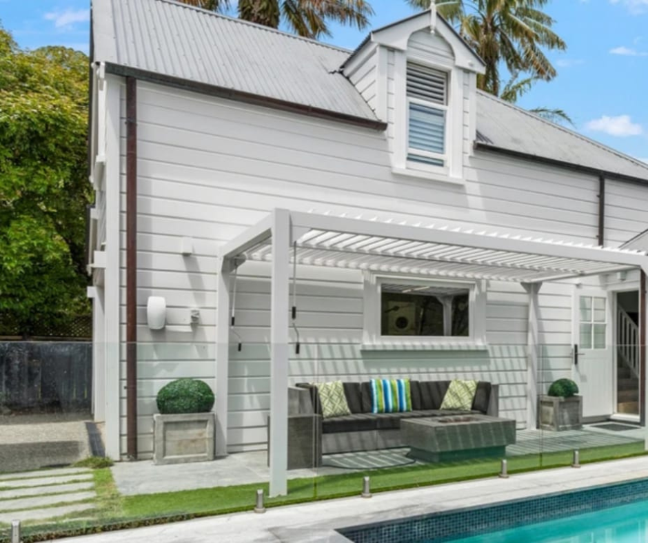 White house with grey roof and a backyard pool, outdoor seating with cushions under a pergola, and palm trees in the background.