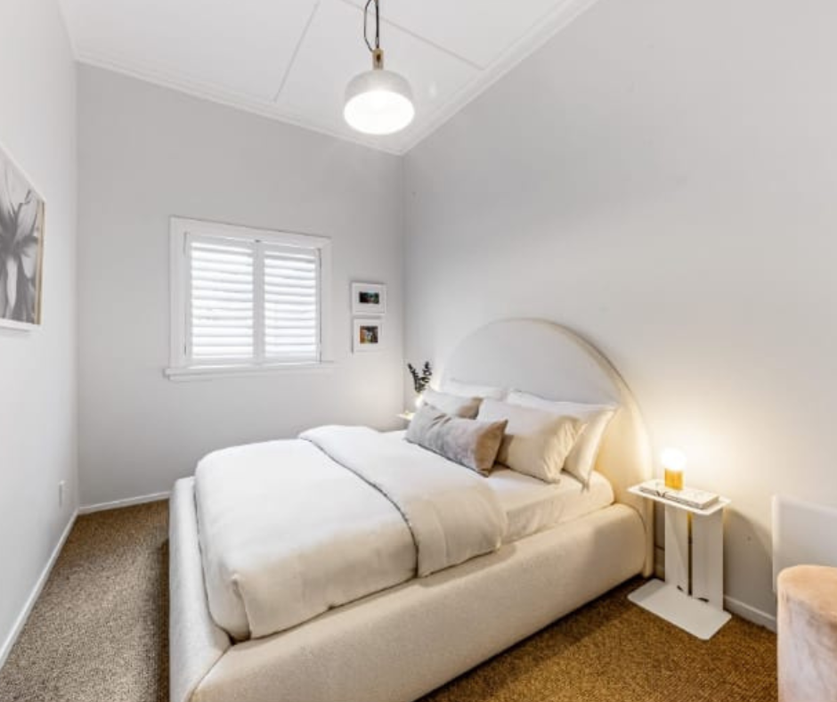 A cozy bedroom with a white bed, pillows, a nightstand with a lamp, and a window with white blinds.