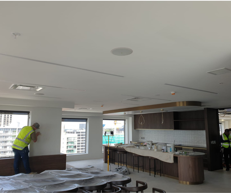 Workers in yellow safety vests working on interior construction in a high-rise apartment or office, with large windows showing city buildings outside.