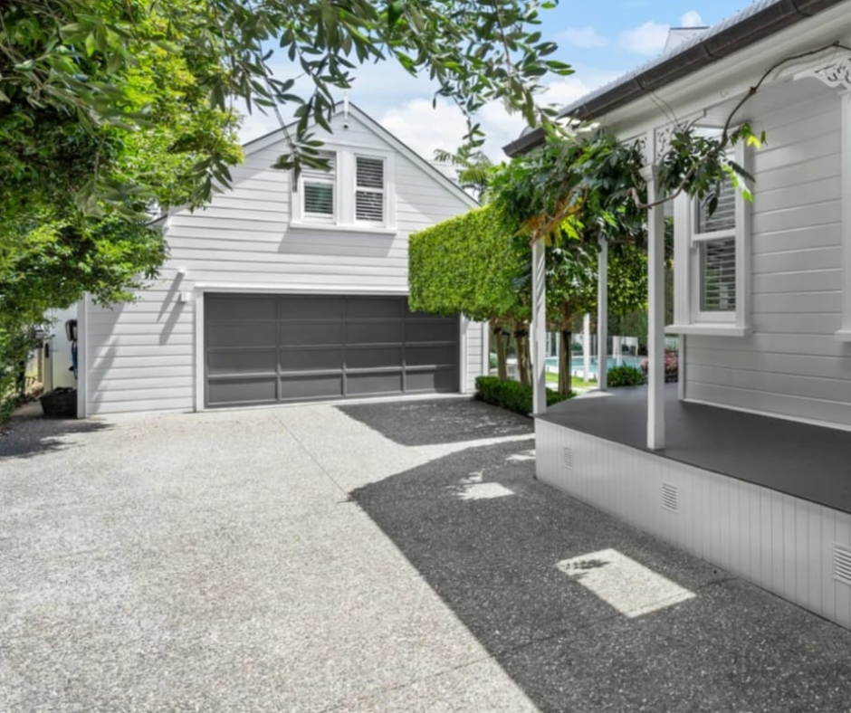 A residential driveway with two houses, trees, and a garage door in the background on a partly cloudy day.