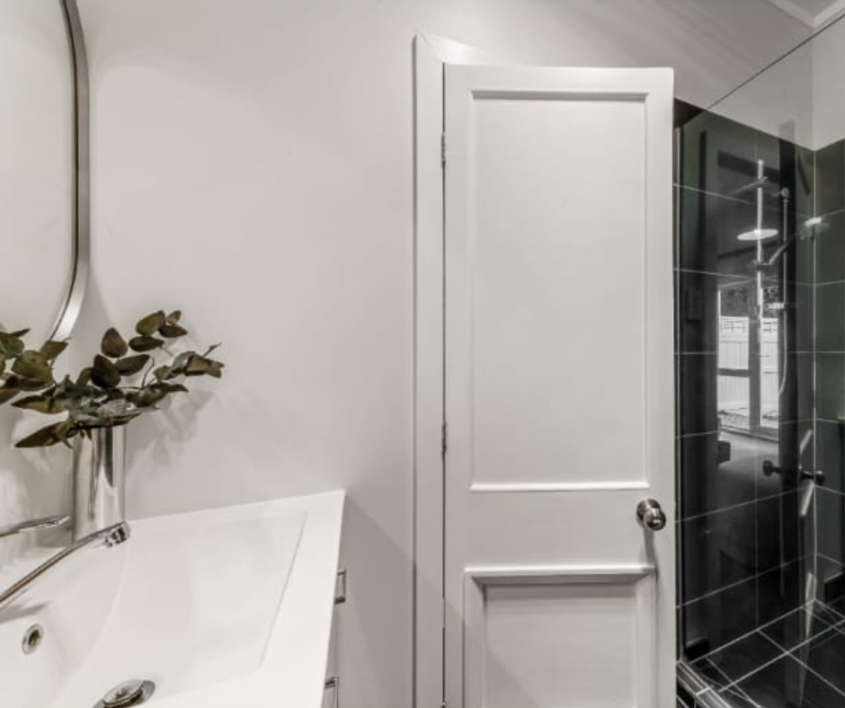 Bathroom with a white door, black tiled shower, a sink with a silver faucet, a mirror, and a vase with dried leaves.