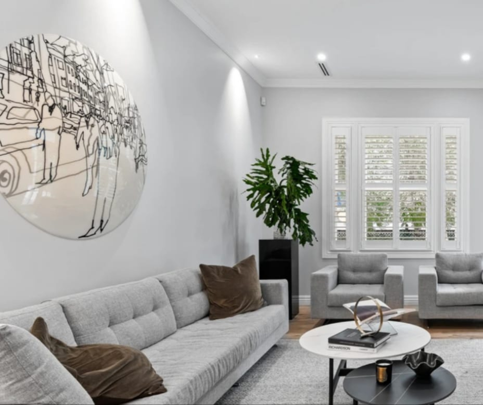 Modern living room with light gray sofa and armchairs, brown pillows, a white round coffee table with books and decor, a tall green houseplant, a large abstract wall art, and plantation shutters on the windows.
