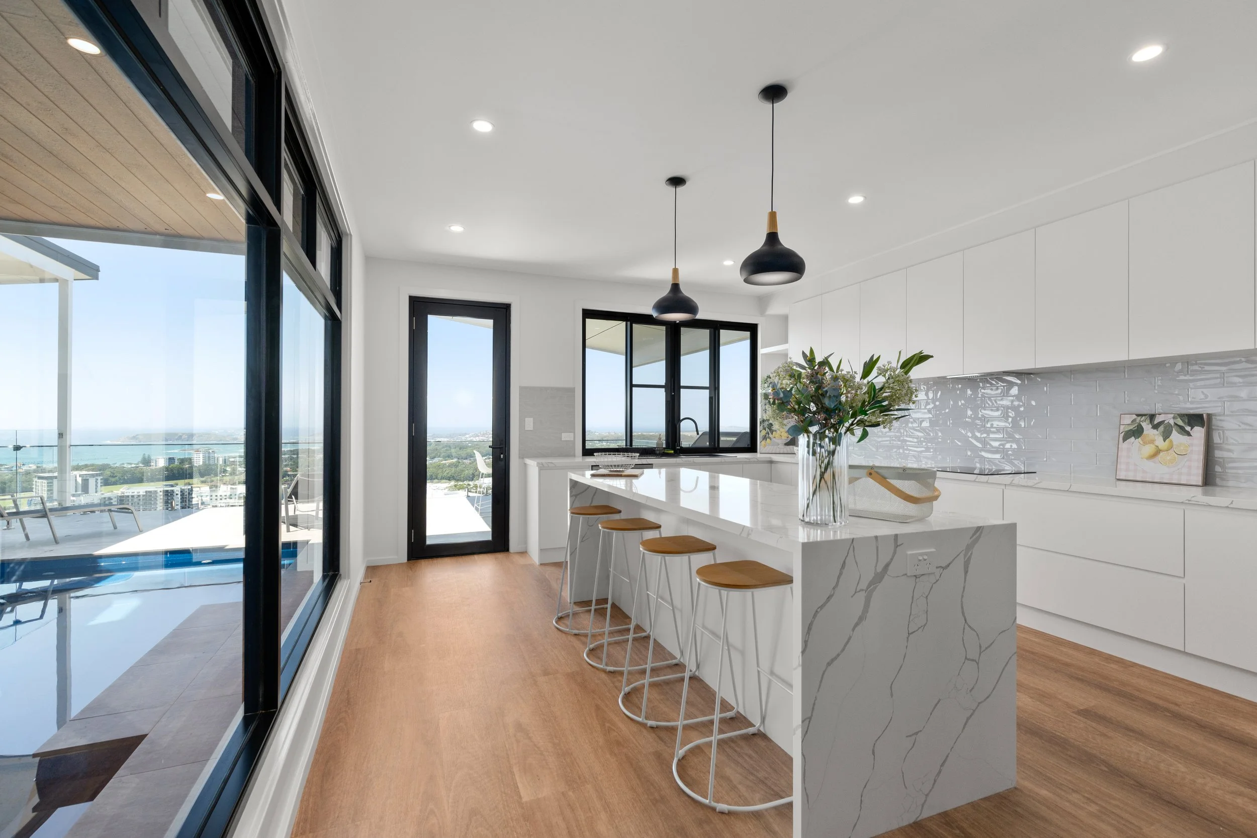 Modern kitchen with white cabinets and marble island, black pendant lights, glass doors and windows, wooden floor, view of city and water outside.