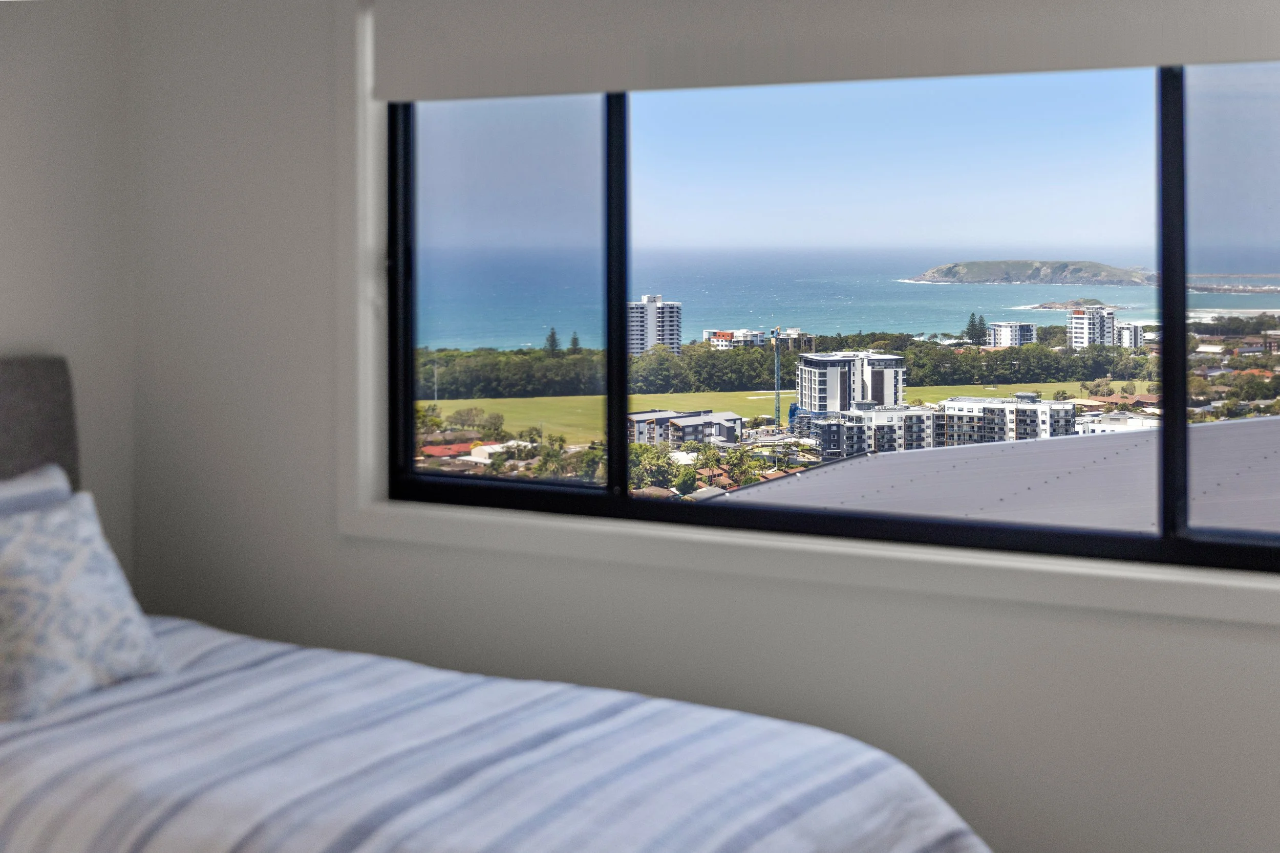 Room with a bed and a large window showing a cityscape with green fields, buildings, and the ocean in the background.