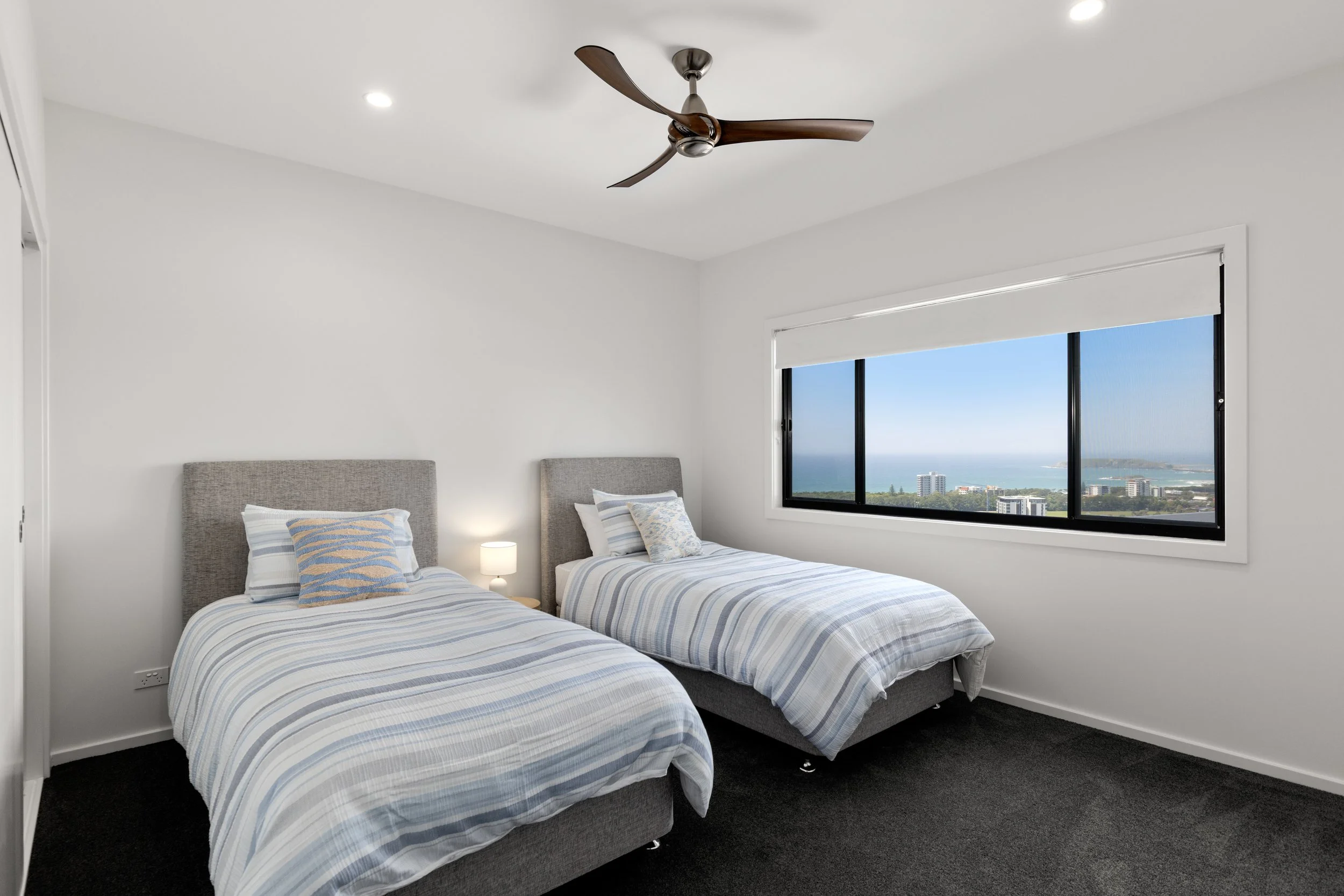 A bedroom with two beds, each with striped bedding and pillows, a nightstand with a lamp between them, a large window with a view of a cityscape and water, white walls, ceiling fan, and carpeted floor.